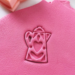 Pode incluir: Um cortador de biscoitos de plástico rosa em forma de cafeteira com corações. O cortador de biscoitos está sobre uma superfície rosa.
