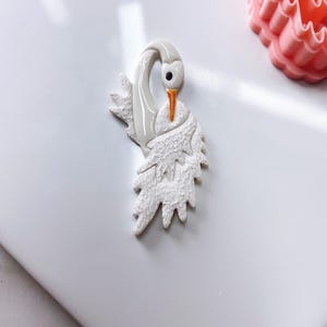 May include: White ceramic swan figurine with a textured surface and orange beak. The swan is in a curled position with its wings spread out.