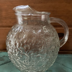 May include: A clear glass pitcher with a textured, frosted surface. The pitcher has a round body and a handle.