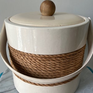 May include: Cream-colored ice bucket with a woven rope design around the middle. The bucket has a matching lid with a wooden knob handle and white handles. A stylish container for entertaining.