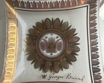 May include: Square glass ashtray with gold and brown accents. The center features a crown design surrounded by a sunburst pattern. The words "Georges Briard" are written in cursive below the crown.