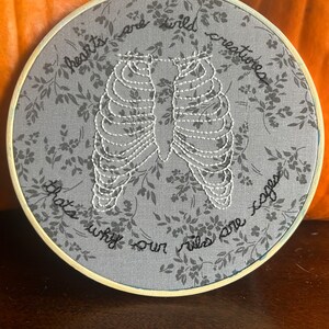 May include: Embroidered hoop art with a gray background and white floral pattern. The hoop features a white embroidered ribcage with the text "hearts are wild creatures that's why our rules are softer".