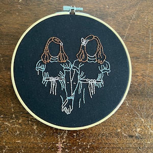 May include: Embroidered hoop art featuring two girls with brown hair and blue dresses holding hands. The girls are outlined in white thread and their hair is brown thread. The background is black fabric.