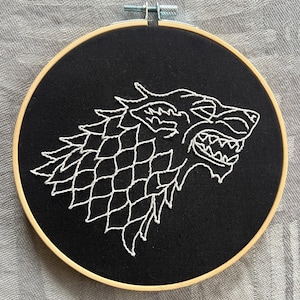 May include: Embroidered white direwolf head on a black fabric background in a wooden embroidery hoop.