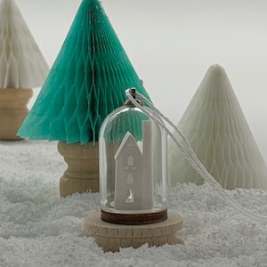 May include: A white paper house ornament inside a clear glass dome with a wooden base. The ornament is sitting on a white surface with two paper trees in the background.