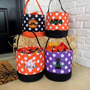 K&ouml;nnte beinhalten: Vier Halloween-Trick-or-Treat-Taschen mit personalisierten Namen. Die Taschen sind orange, lila und schwarz mit wei&szlig;en Punkten. Die Taschen zeigen einen K&uuml;rbis, eine Fledermaus, ein Gespenst und eine Katze.