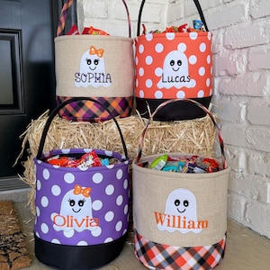 May include: Four cylindrical fabric buckets for collecting candy, each featuring a ghost applique and a personalized name. The buckets display various patterns including orange polka dots, purple polka dots, and plaid, with black handles and bases.