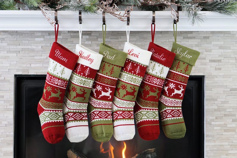 May include: Six personalized Christmas stockings hanging in a row. Each stocking features a knit pattern with reindeer, snowflakes, and geometric shapes in red, white, and olive green. The names Mom, Hadley, Dad, Levi, Natalie, and Skylar are embroidered.