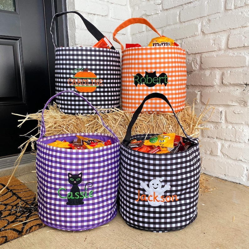May include: Four Halloween trick-or-treat bags with personalized names. The bags are made of black and white gingham fabric and feature different Halloween-themed designs. The names on the bags are "Cassie", "Robert", "Jackson", and "Caroline".