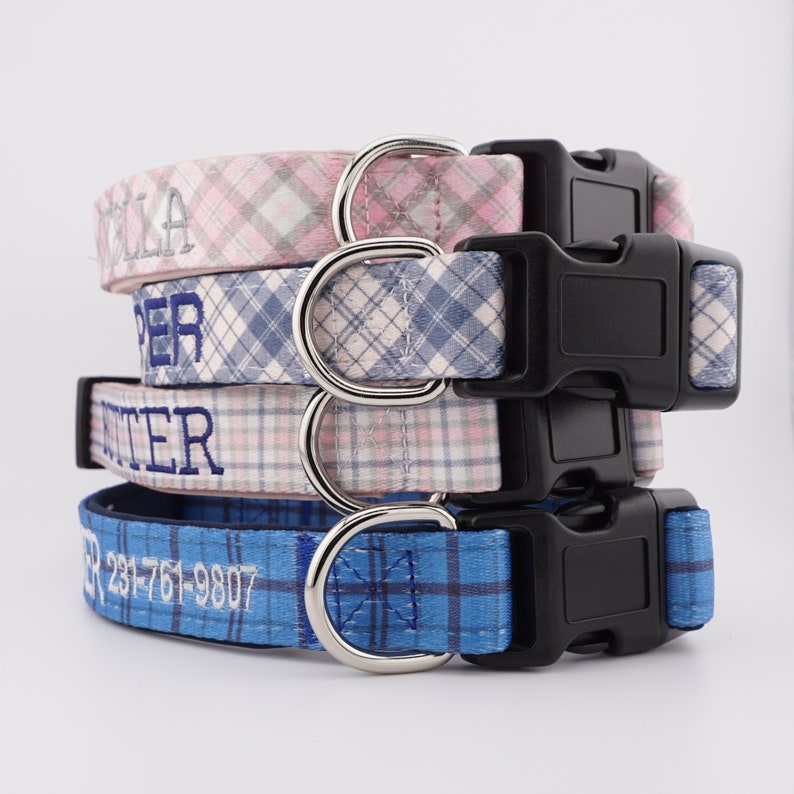 May include: A stack of four fabric dog collars featuring plaid patterns in pink, blue, and white. Each collar includes a black plastic side-release buckle, a silver metal D-ring, and custom embroidery. The bottom collar displays the text 231-761-9807.
