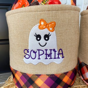 May include: A burlap trick-or-treat bag with a black and orange plaid trim. The bag features an embroidered white ghost with a pink bow and the name "Sophia" in purple.