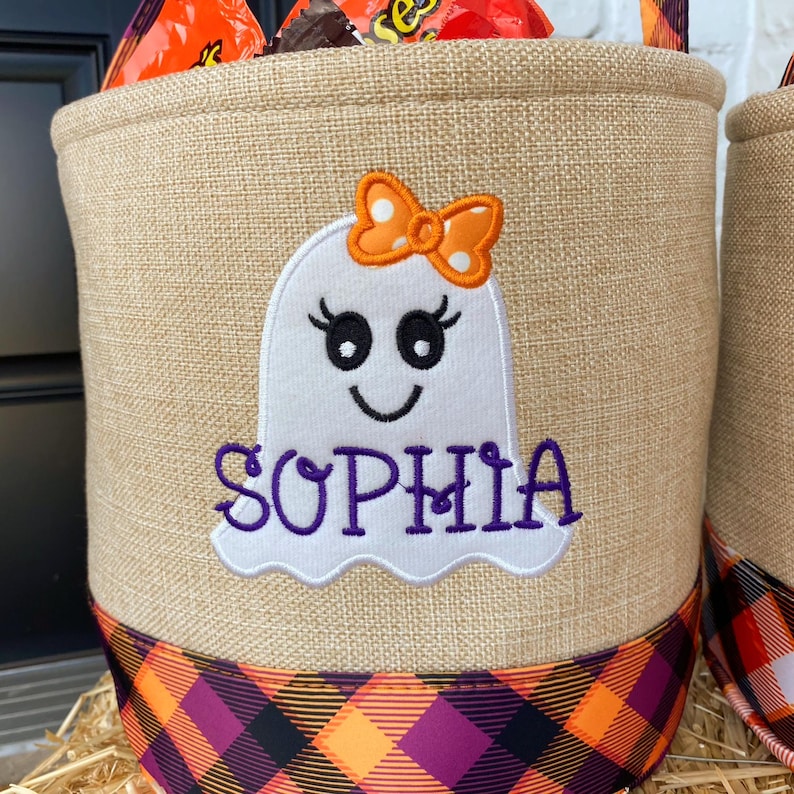 May include: A burlap trick-or-treat bag with a black and orange plaid trim. The bag features an embroidered white ghost with a pink bow and the name "Sophia" in purple.