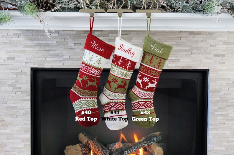 May include: Three knit Christmas stockings featuring reindeer, snowflake, and tree patterns in red, white, and green. The stockings are labeled #40 Red Top, #41 White Top, and #42 Green Top, with embroidered names Mom, Hadley, and Dad on the cuffs.