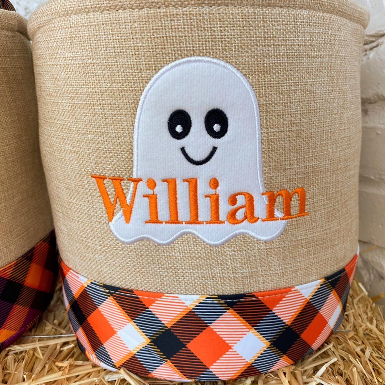 May include: A burlap storage bin with a black and orange plaid trim. The bin features an embroidered white ghost with black eyes and a smile, and the name "William" in orange lettering.