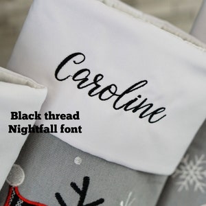 May include: A gray Christmas stocking with a white cuff featuring the name Caroline embroidered in black thread using Nightfall font. The stocking is decorated with embroidered snowflakes and a partial graphic design.