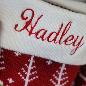 May include: A red and white knit Christmas stocking featuring a white cuff embroidered with the name Hadley in red script. The stocking body displays a repeating pattern of white pine trees against a deep red background.