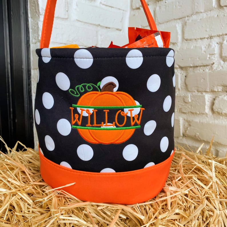 K&ouml;nnte beinhalten: Ein schwarz-wei&szlig;er Tupfen-Trick-or-Treat-Eimer mit einem orangefarbenen K&uuml;rbis und dem Namen "WILLOW", der darauf gestickt ist. Der Eimer hat einen orangefarbenen Rand.