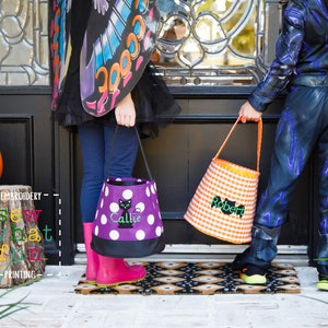 May include: Two children dressed in Halloween costumes are holding trick-or-treat bags. The girl is wearing a butterfly costume and a purple bag with white polka dots and a black cat embroidered on it. The boy is wearing a black and blue costume and an orange and white checkered bag with the name "Robert" embroidered on it.