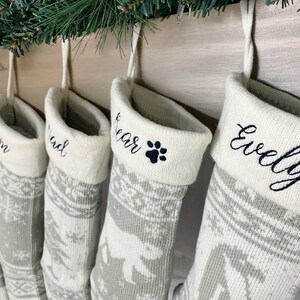 May include: Five personalized Christmas stockings with a white background and gray snowflake pattern. The stockings are hung on a wooden mantel and are embroidered with the names "Evelyn", "Bear", "Chad", "Tom", and "Jon".