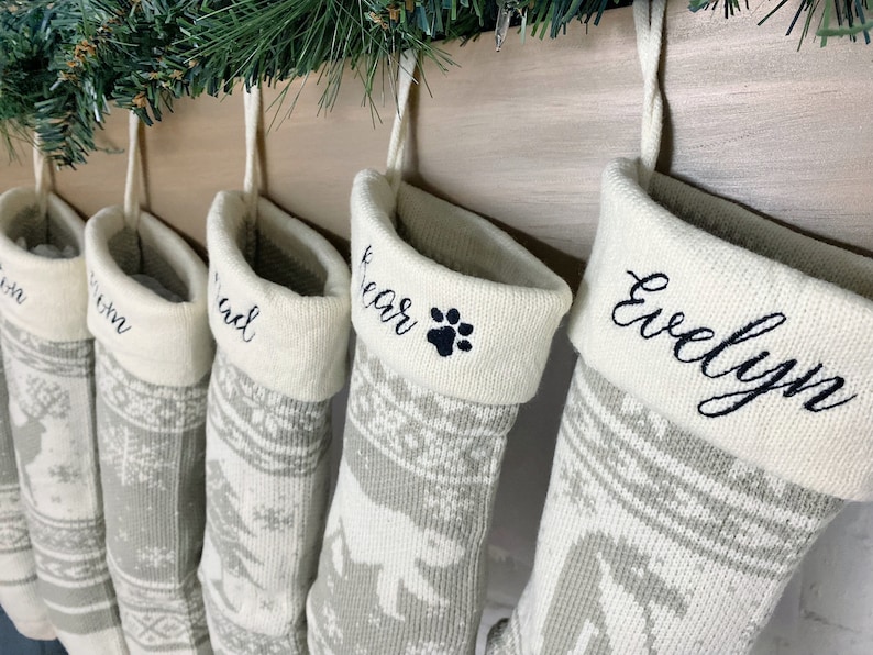 May include: Five personalized Christmas stockings with a white background and gray snowflake pattern. The stockings are hung on a wooden mantel and are embroidered with the names "Evelyn", "Bear", "Chad", "Tom", and "Jon".