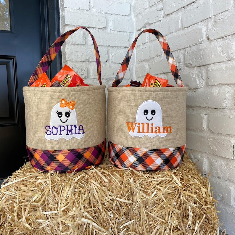 May include: Two personalized Halloween trick-or-treat bags with ghost designs. The bags are made of burlap and have orange and black plaid trim. The bags are filled with candy and have the names "Sophia" and "William" embroidered on them.