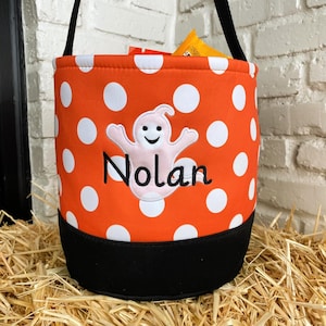 K&ouml;nnte beinhalten: Orangefarbener und wei&szlig;er, gepunkteter S&uuml;&szlig;igkeiteneimer mit schwarzem Boden. Der Eimer hat einen Geist mit einem l&auml;chelnden Gesicht und den Namen "Nolan" bestickt.