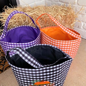 May include: Three Halloween trick-or-treating bags with gingham patterns. The bags are purple, orange, and black and white. The bags have a single handle and are made of fabric.