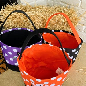 K&ouml;nnte beinhalten: Drei Halloween-Trick-or-Treat-Taschen mit schwarzen und wei&szlig;en Tupfenmustern. Die Taschen sind aus Stoff und haben schwarze Henkel. Die Taschen sind mit orangefarbenem und violettem Stoff gef&uuml;ttert. Eine Tasche hat ein schwarzes Katzenmotiv, eine andere ein Geistermotiv und die letzte ein K&uuml;rbismotiv.