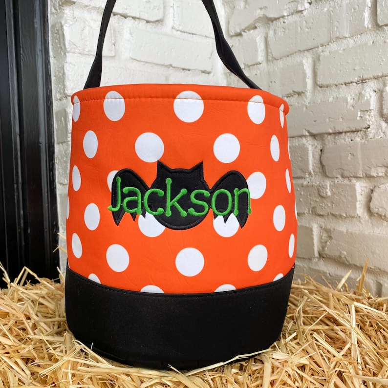 K&ouml;nnte beinhalten: Orangefarbene und wei&szlig;e Tupfen-Trick-or-Treat-Tasche mit einer schwarzen Fledermaus und dem Namen Jackson in Gr&uuml;n gestickt.