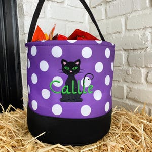 K&ouml;nnte beinhalten: Lilafarbener und wei&szlig;er Tupfen-Trick-or-Treat-Eimer mit einer schwarzen Katze und dem Namen Callie, der auf der Vorderseite gestickt ist.