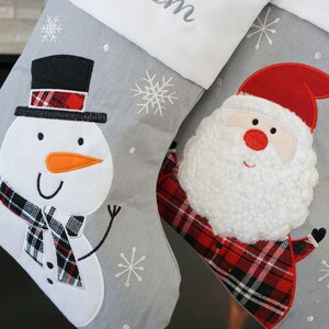 May include: Two gray Christmas stockings featuring white cuffs and embroidered snowflake patterns. One stocking displays a snowman with a black top hat and plaid scarf, while the other features a Santa Claus with a textured beard and plaid coat.