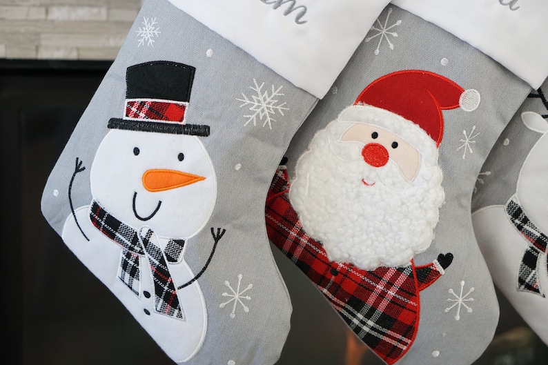 May include: Two gray Christmas stockings featuring white cuffs and embroidered snowflake patterns. One stocking displays a snowman with a black top hat and plaid scarf, while the other features a Santa Claus with a textured beard and plaid coat.
