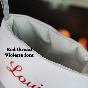 May include: A white Christmas stocking featuring the text Red thread Violetta font embroidered in red. The stocking has a light gray interior lining and a matching gray hanging loop at the top, with a partial view of a name starting with Loui.
