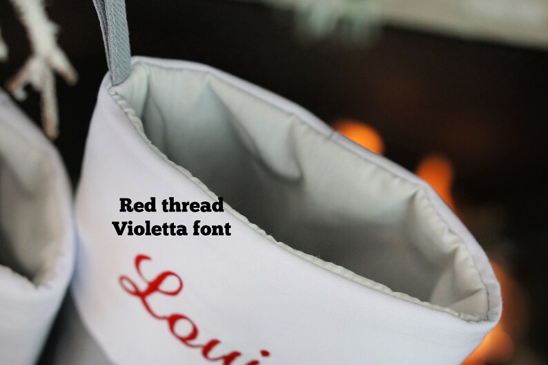 May include: A white Christmas stocking featuring the text Red thread Violetta font embroidered in red. The stocking has a light gray interior lining and a matching gray hanging loop at the top, with a partial view of a name starting with Loui.