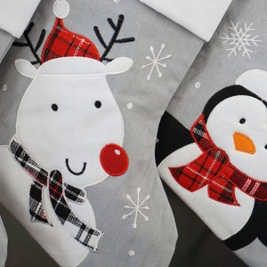 May include: A set of gray Christmas stockings featuring embroidered appliqu&eacute;s of a reindeer with a red nose and plaid hat, and a penguin wearing a red Santa hat and a red plaid scarf, accented with white stitched snowflake patterns.