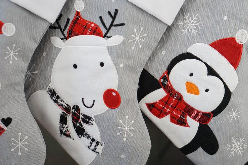 May include: A set of gray Christmas stockings featuring embroidered appliqu&eacute;s of a reindeer with a red nose and plaid hat, and a penguin wearing a red Santa hat and a red plaid scarf, accented with white stitched snowflake patterns.
