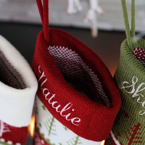 May include: Three knit Christmas stockings featuring a festive tree pattern. The stockings are white, red, and olive green, each with a hanging loop. The cuffs are personalized with the embroidered names Levi, Natalie, and Skylar in script.