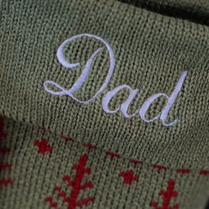 May include: An olive green knitted Christmas stocking featuring a folded cuff embroidered with the word Dad in white script lettering. The body of the stocking displays a repeating pattern of small red pine trees and geometric star shapes.