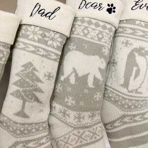 May include: Four white and gray knitted Christmas stockings with personalized names: "Mom", "Dad", "Bear", and "Evelyn". Each stocking features a different winter-themed design, including a pine tree, a polar bear, and a penguin.