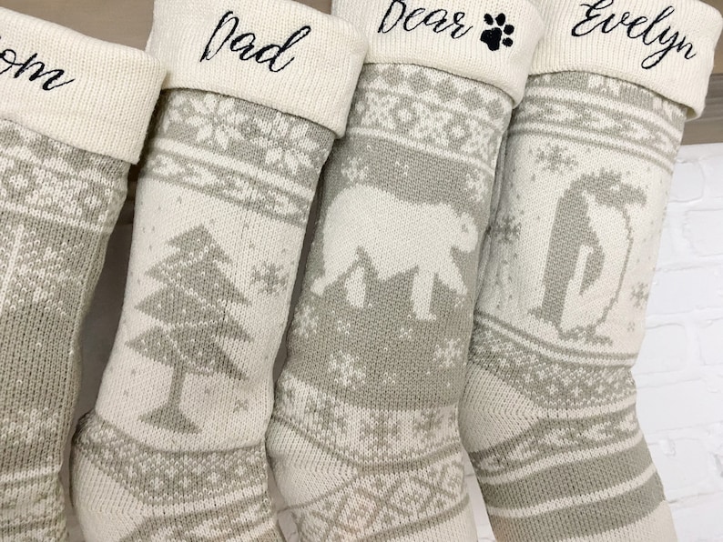 May include: Four white and gray knitted Christmas stockings with personalized names: "Mom", "Dad", "Bear", and "Evelyn". Each stocking features a different winter-themed design, including a pine tree, a polar bear, and a penguin.
