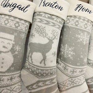 May include: Four gray and white knitted Christmas stockings with different designs. The stockings are personalized with the names Abigail, Trenton, Mom, and a fourth name that is not visible.