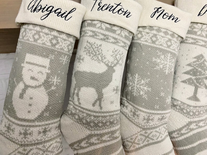 May include: Four gray and white knitted Christmas stockings with different designs. The stockings are personalized with the names Abigail, Trenton, Mom, and a fourth name that is not visible.