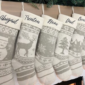 May include: Six white and gray knitted Christmas stockings with personalized names: Abigail, Trenton, Mom, Dad, Bear, and Eve. Each stocking features a different winter-themed design, including a snowman, a deer, snowflakes, a pine tree, a cabin, and a Christmas tree.