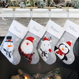 May include: Four gray Christmas stockings with white cuffs embroidered with the names Mom, Dad, Harper, and William. Each stocking features a festive character: a snowman, Santa Claus, a reindeer, and a penguin, all wearing red plaid accents.