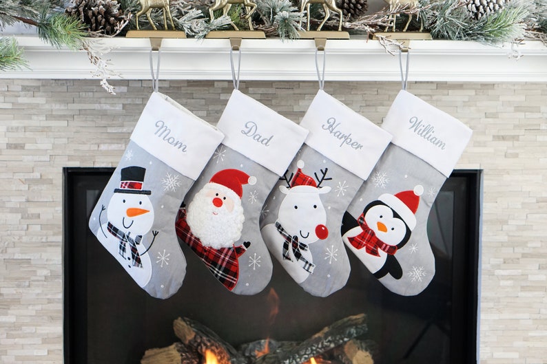 May include: Four gray Christmas stockings with white cuffs embroidered with the names Mom, Dad, Harper, and William. Each stocking features a festive character: a snowman, Santa Claus, a reindeer, and a penguin, all wearing red plaid accents.