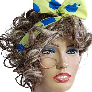 May include: A yellow headband with blue apple prints tied in a bow. The headband is worn on a mannequin with curly brown hair.