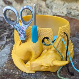 May include: A yellow dragon-shaped yarn bowl with a teal crochet hook and floral scissors sticking out of the top.  The bowl is holding three skeins of yarn in green, blue, and black.