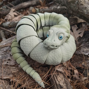 May include: A light gray, segmented, snake-like sculpture with blue eyes. The sculpture is made of a material that resembles scales and is coiled in a circle.