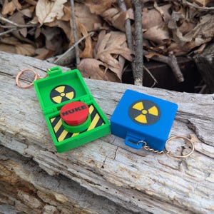 May include: Two keychains shaped like small boxes with a nuclear symbol and a red button labeled "NUKE". One keychain is green and the other is blue.
