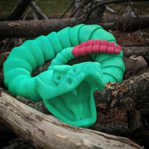 May include: A green and red plastic snake toy with an open mouth, lying on a bed of brown twigs and pine needles.